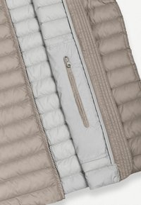 Közelkép egy bézs és szürke steppelt dzsekiről, amely bemutatja a belső bélést, egy függőleges cipzáras zsebet és a varrási részleteket.