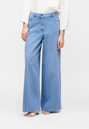 Frau trägt hellblaue, hochgeschnittene, weit geschnittene Jeans mit dünnen weißen Vertikalstreifen und weißen offenen Sandaletten, steht mit entspannten Händen.