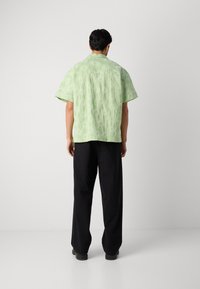 Short-sleeved, light green shirt with a sunburst pattern, paired with loose black trousers and black shoes. Minimalistic design, relaxed fit.