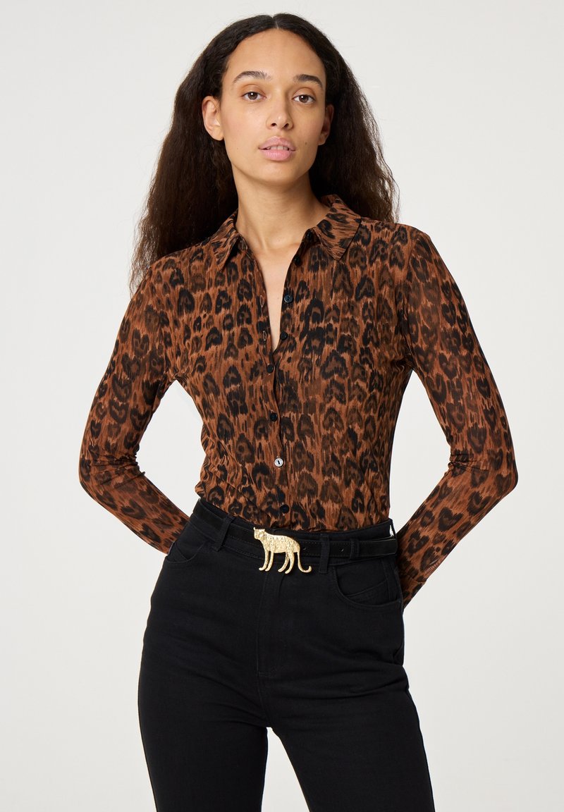 Braunes Langarmshirt mit Leopardenmuster, spitzem Kragen und Knopfleiste, kombiniert mit hoch taillierten schwarzen Jeans und einer goldenen Gürtelschnalle in Tierform.