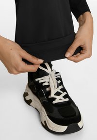 Black stretchy pants with a smooth finish, featuring a snug hem, paired with a black sneaker with white laces and contrasting sole accents.