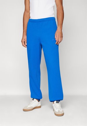 Pantalons de survêtement bleus avec une coupe ample, une taille élastique et un détail de logo. Assortis avec des baskets blanches et un débardeur blanc ajusté.