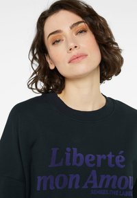 Schwarzer Sweatshirt mit rundem Ausschnitt und lila Text "Liberté mon Amour", der prominent auf der Vorderseite platziert ist. Glatte Textur.