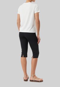 Biała bawełniana koszulka, dopasowane czarne spodnie capri z bocznymi rozcięciami oraz sandały na cienkich paskach w kolorze złotym. Widok z tyłu, podkreślający szczegóły stroju.