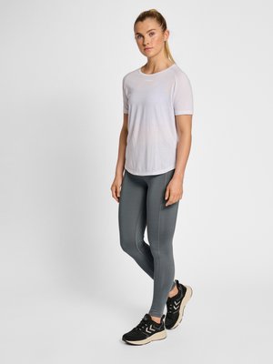 Vrouw staand in een wit T-shirt met korte mouwen, grijze legging en zwarte sneakers tegen een effen lichte achtergrond.
