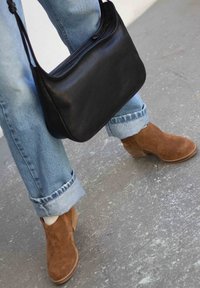 Stivaletti in suede marrone con tacco a blocco, indossati con jeans azzurri arrotolati. Una borsa a tracolla in pelle nera è posizionata nelle vicinanze.