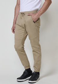 Pantaloni jogger bej cu talie elastică, buzunare laterale și detalii de cusătură la genunchi, asortați cu adidași negri și șosete cu model.