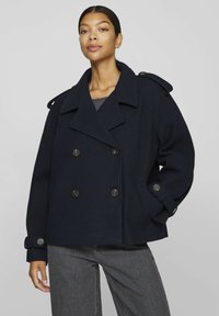 Mörkblå ull pea coat med dubbelknäppt front, stora slag, axelklaffar och knäppte ärmslut, kombinerad med grå denim.