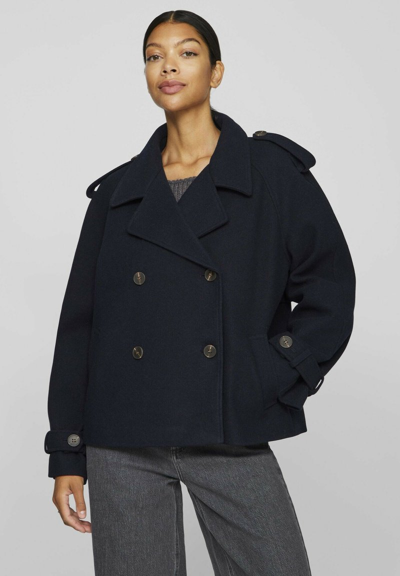 Mörkblå ull pea coat med dubbelknäppt front, stora slag, axelklaffar och knäppte ärmslut, kombinerad med grå denim.