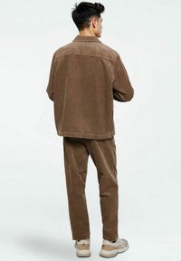 Veste en velours côtelé marron avec un pantalon assorti, présentant une texture à côtes verticales, un col boutonné et des baskets de couleur neutre.