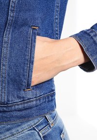 Džínová bunda s přiléhavým střihem, modré barvy a viditelným prošíváním. Ruka je umístěna v boční kapse, což ukazuje umístění kapsy.