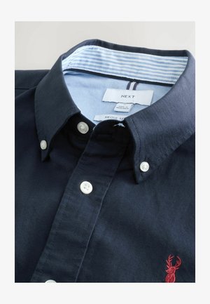 Camicia blu navy con bottoni in cotone, caratterizzata da una superficie testurizzata, bottoni bianchi e un colletto a righe azzurre con una piccola logo rosso.