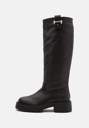 Bota alta de cuero negro con suela gruesa y detalle de correa con hebilla plateada cerca de la parte superior sobre un fondo claro y liso.