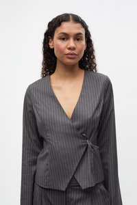 Mladá žena s kučeravými vlasmi, oblečená v sivom prúžkovanom zavinovacom saku viazanom v páse, stojaca pred jednoduchým bielym pozadím.