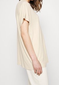 Beige topp med korta ärmar i texturerat tyg med lös passform och subtila vertikala ränder, visad från sidan.