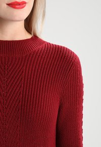 Roter gerippter Strickpullover mit hohem Kragen, der ein vertikales Muster und einen strukturierten Stoff aufweist. Nahaufnahme des Schulter- und Kragenbereichs.
