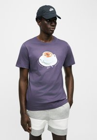 Νέος άνδρας που φοράει μαύρο καπέλο Nike, μωβ μπλούζα με σχέδιο κούπας καφέ και γκρι σορτς με λευκές επιφάνειες, στέκεται με το ένα χέρι στην τσέπη.