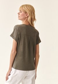 Olivgrünes Kurzarmoberteil mit Rundhalsausschnitt und einer Rückenfalten-Detail, kombiniert mit weißen Leinenhosen. Weiche Textur, lässiges Design.