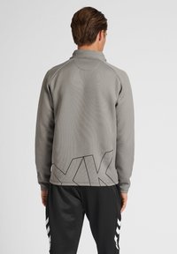Felpa grigia sportiva con maniche lunghe e colletto alto, caratterizzata da un logo geometrico nero sul retro e un tessuto testurizzato.