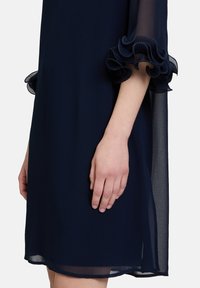 Robe bleu marine avec un tissu transparent, des accents froncés aux manches et une coupe ample. L'ourlet est légèrement arrondi pour permettre le mouvement.