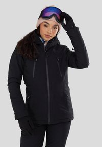 Vrouw in een zwarte winterjas en handschoenen die paarse skibril boven een roze muts afstelt, staand tegen een lichtgrijze achtergrond.