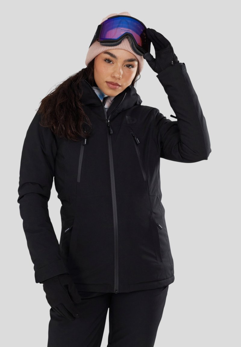 Vrouw in een zwarte winterjas en handschoenen die paarse skibril boven een roze muts afstelt, staand tegen een lichtgrijze achtergrond.