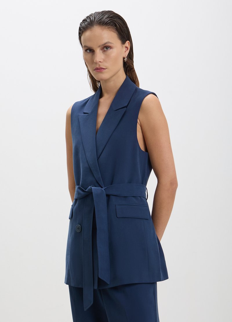 Gilet sans manches bleu marine en tissu lisse, avec un décolleté en V profond, une taille ceinturée et des poches latérales. Coupe ajustée avec une silhouette structurée.