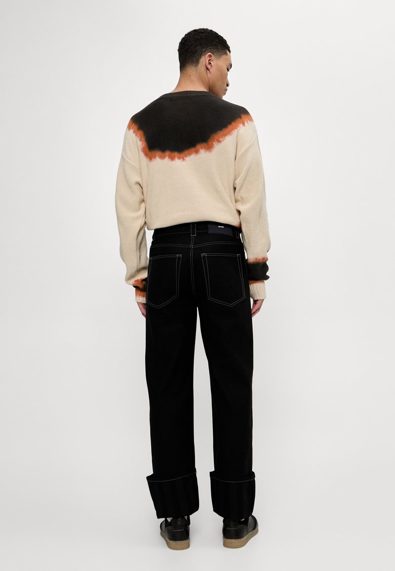 Beige Rundhalspullover mit schwarzem und orangefarbenem Dip-Dye-Effekt, kombiniert mit schwarzen weit geschnittenen Jeans und schwarzen Schuhen, die einen umgeschlagenen Bund haben.