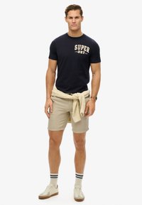 Camiseta de algodón navy con estampado "SUPER DRY", pantalones cortos beige, suéter de punto atado a la cintura, zapatillas blancas y calcetines a rayas.