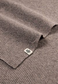Brown ribbed knit fabric with a textured surface, featuring a small square label with a logo at the edge.