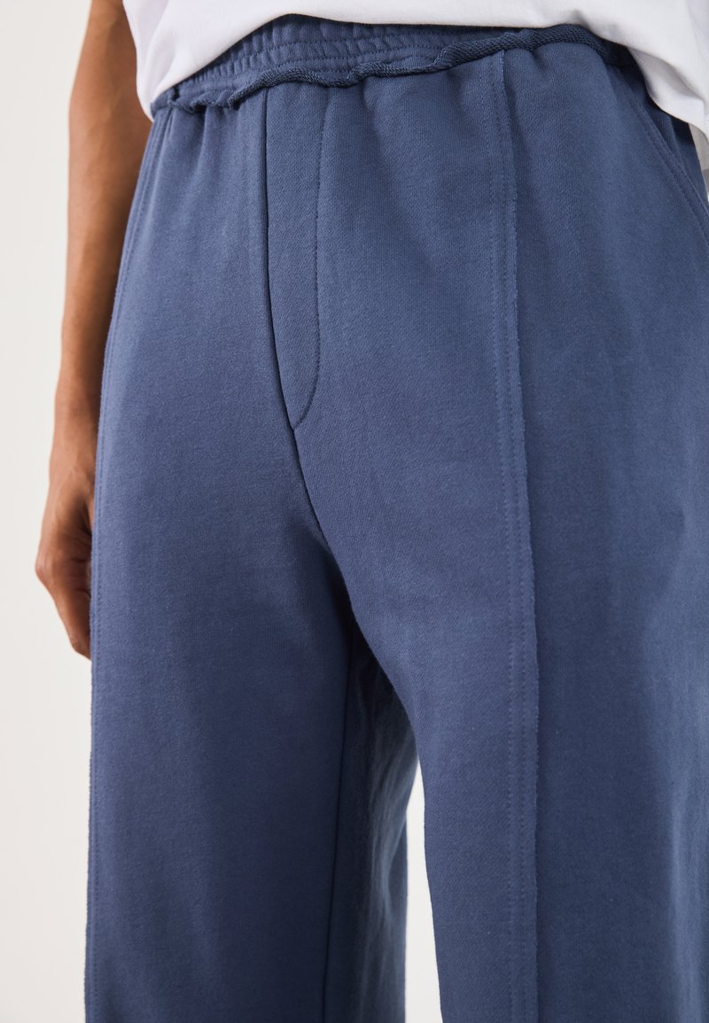 Pantaloni della tuta blu navy realizzati in tessuto morbido, caratterizzati da una cintura larga, tasche laterali e cuciture visibili lungo le giunture.
