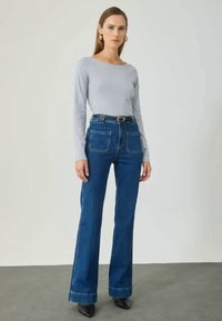 Lys grå langærmet top parret med højtaljede blå flagrende jeans. Jeansene har to baglommer og en denimtekstur. Sorte hælede støvler.