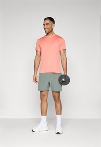 Tricou sportiv din coral ușor, pantaloni scurți verzi, adidași albi, ținând o disc de greutate neagră, stând pe un fundal neutru.
