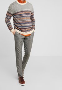 Maglione lavorato a maglia con motivi a strisce in grigio, blu e arancione; pantaloni a quadretti grigi; sneakers stringate marroni; design minimalista.