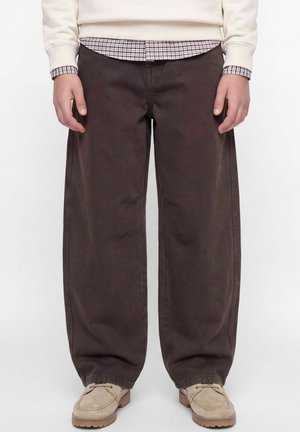 Personne portant un pantalon ample marron foncé, des chaussures beiges, un pull crème et une chemise à carreaux en dessous, debout avec les mains détendues.