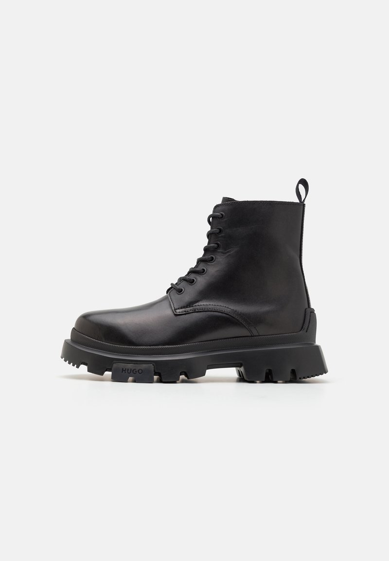 Botte montante en cuir noir avec semelle à crampons épaisse et languette à l'arrière, marquée "HUGO" sur le côté de la semelle, vue de profil sur fond blanc.
