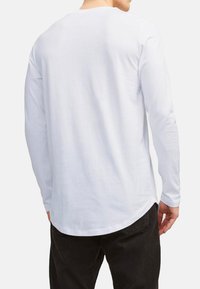 Chemise blanche à manches longues avec un ourlet arrondi, fabriquée en tissu doux. Présente un col rond et des coutures d'épaule basses. Pas de logos visibles.
