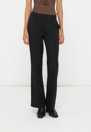 Femme portant un pantalon noir droit avec passants de ceinture, associé à des chaussures à talons noires et un haut marron sans manches.