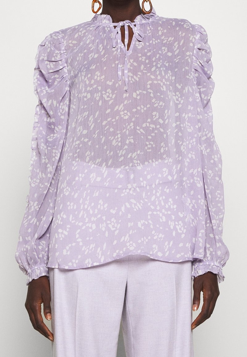 Femme portant un chemisier lavande transparent avec un motif blanc tacheté, des épaules à volants, un col à nouer et de longues manches bouffantes avec des poignets froncés.