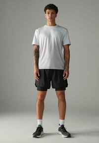 Lysgymnastiktrøje i grå med rund hals og korte ærmer, parret med sorte shorts. Modellen står i sorte sneakers med hvide detaljer.
