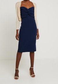 Tmavě modré šaty s přiléhavým střihem, láskyplným výstřihem a šroubovaným detailem na látce. Doplněné béžovým kardiganem a tělovými vysokými podpatky.