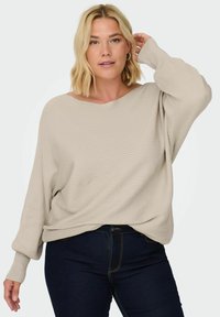 Beige ribbed sweater with a wide neckline, long sleeves, and relaxed fit, paired with dark blue jeans.