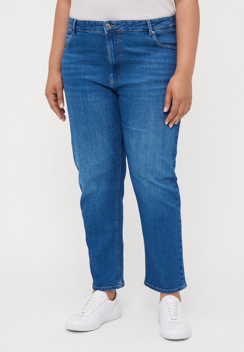 Jeans de ganga azul com corte reto, apresentando fecho de botão, passadores para cinto e um ligeiro desgaste. Combinados com ténis brancos.