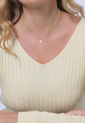 Gold beschichtete Halsketten mit floralen Akzenten und einem runden Anhänger, getragen über einem hellbeigen gerippten Oberteil mit V-Ausschnitt.