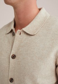 Pulover tricotat bej cu guler, având nasturi mari maro și un material texturat. Detaliile de prim-plan pun în evidență cusăturile.