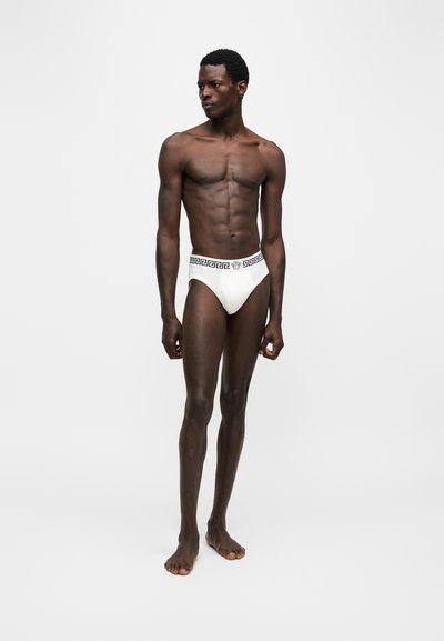 Mannequin homme debout en slip blanc avec ceinture noire à motif grec sur fond blanc uni, regardant vers la gauche.