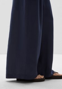 Široke hlače iz temno modrega blaga z gladko teksturo. Imajo sproščen kroj in so v kombinaciji z rjavimi flat sandals.