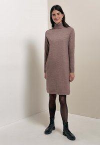Femme portant une robe-pull à col roulé mauve, des collants noirs transparents et des bottines noires à lacets, debout devant un fond neutre.
