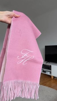 Ροζ μάλλινο κασκόλ με φουντωτές άκρες, διακοσμημένο με λευκά κεντημένα αρχικά "FH" στη μία πλευρά. Μαλακή υφή, ορθογώνιο σχήμα.