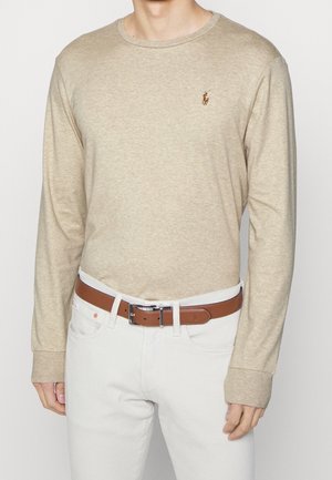 Chemise beige à manches longues avec un petit logo sur la poitrine, rentrée dans un pantalon blanc avec une ceinture en cuir marron.